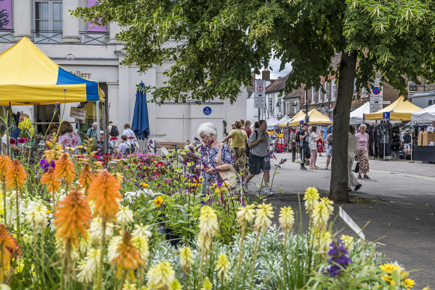 Colourful flowers in the foreground of St Albans Market, with shoppers, yellow market canopies and the city centre streets unfolding behind them.